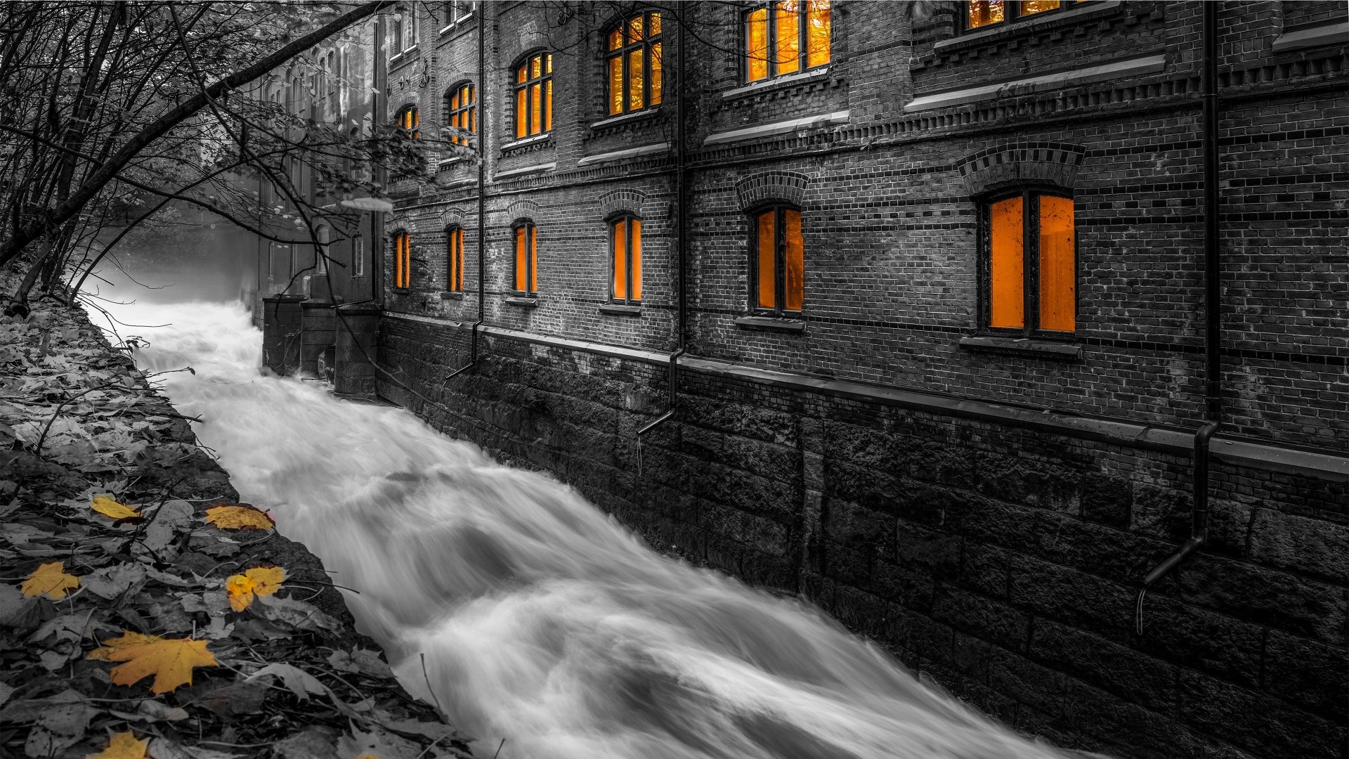 4K Ultra HD wallpaper of a man-made brick building beside a rushing stream, with glowing orange windows contrasting the black and white surroundings.