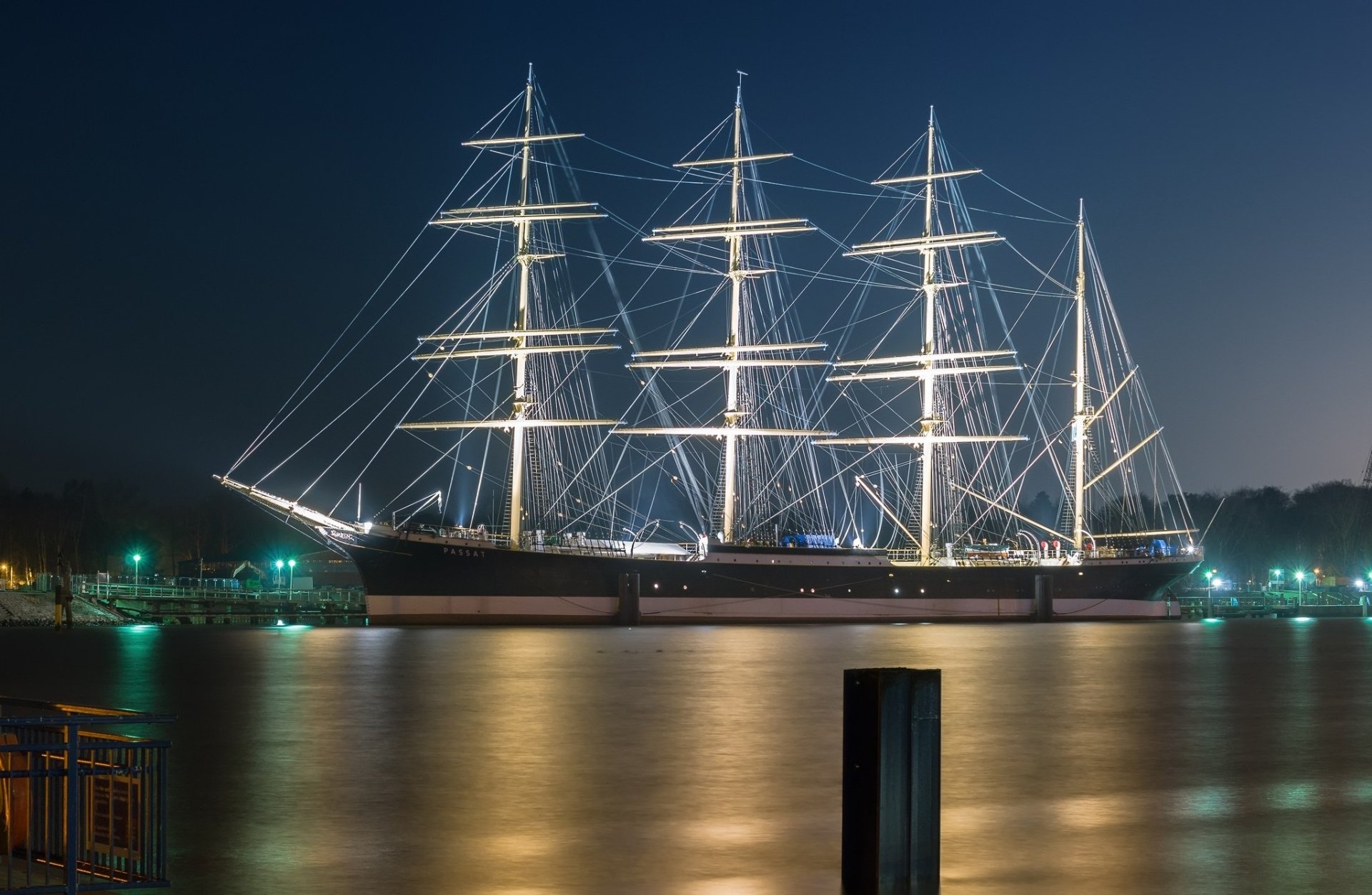 A majestic barque sailboat illuminated at night, reflected in calm waters with a faint outline of a Volkswagen Passat nearby, captured in an HD desktop wallpaper.