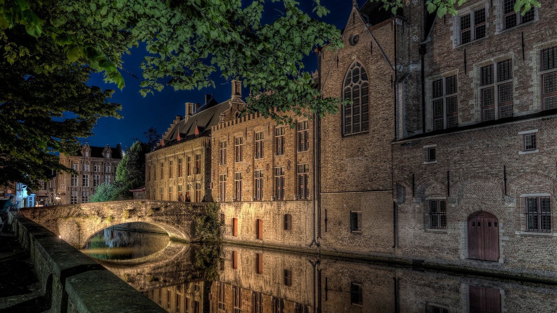 HD desktop wallpaper showing a historic house and stone bridge over a calm canal, illuminated at night with tree branches framing the scene.