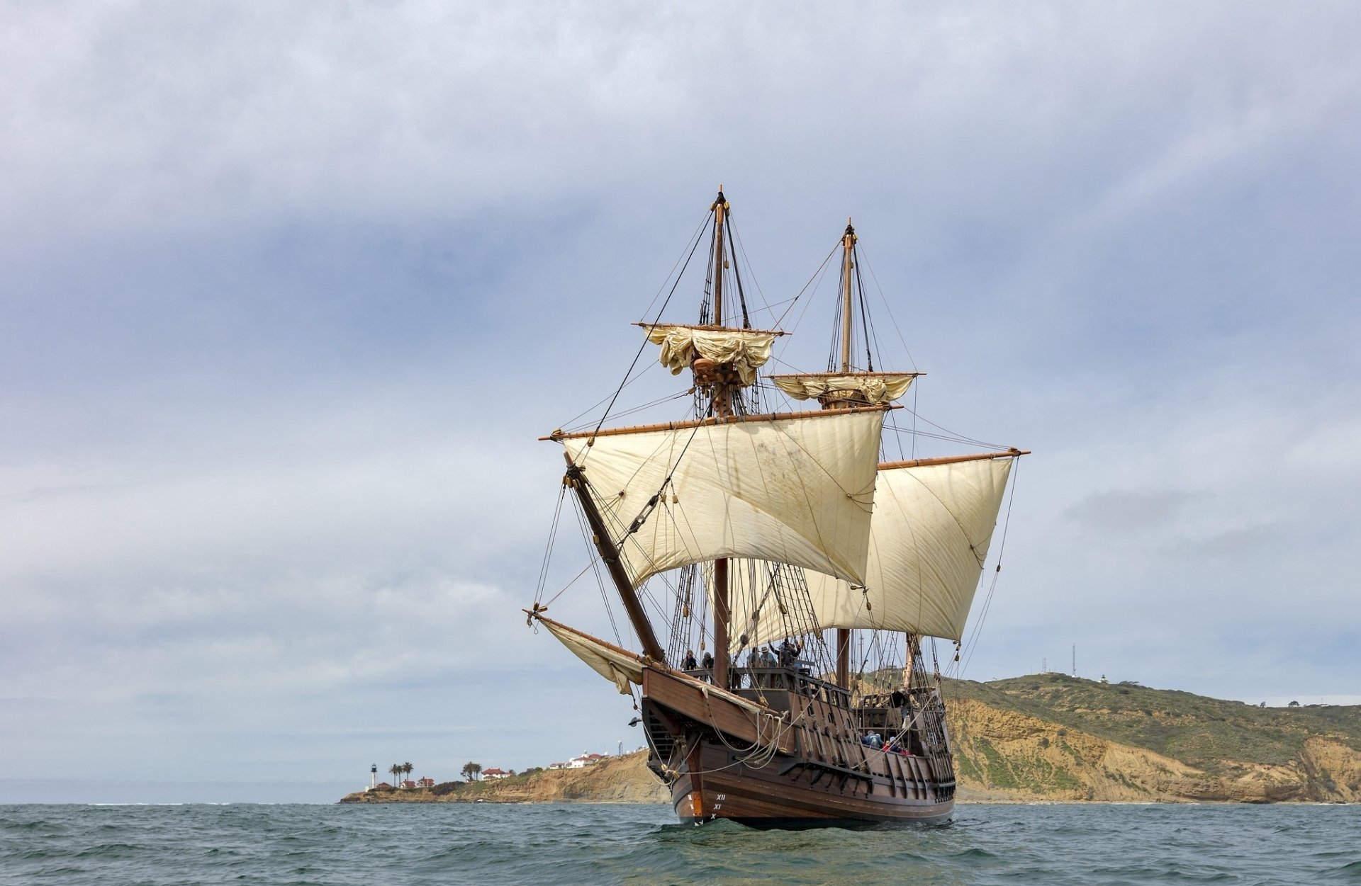 HD desktop wallpaper showing a sailboat sailing on the sea with sails unfurled under a cloudy sky near a distant shoreline.