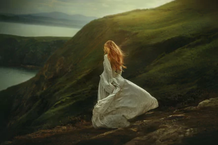 Rear view of a woman in a flowing white dress standing on a windswept grassy cliff overlooking the sea, moody light — 2K Quad HD PC desktop wallpaper/background.