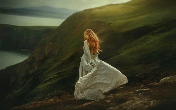 Rear view of a woman in a flowing white dress standing on a windswept grassy cliff overlooking the sea, moody light — 2K Quad HD PC desktop wallpaper/background.