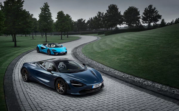 HD desktop wallpaper featuring two McLaren supercars, a dark blue 720S and a blue 570S Spider, on a winding cobblestone road surrounded by greenery and trees.
