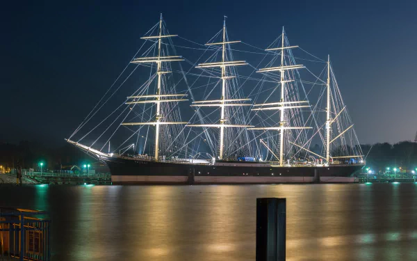 A majestic barque sailboat illuminated at night, reflected in calm waters with a faint outline of a Volkswagen Passat nearby, captured in an HD desktop wallpaper.