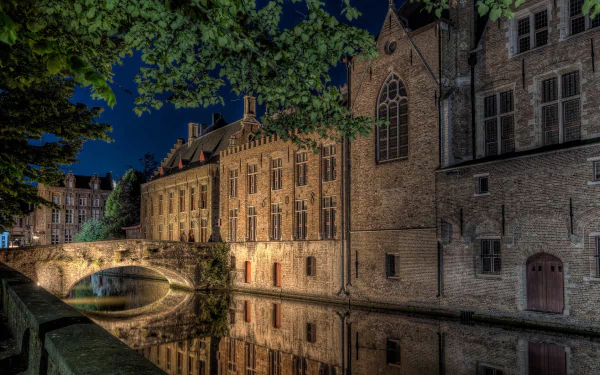 HD desktop wallpaper showing a historic house and stone bridge over a calm canal, illuminated at night with tree branches framing the scene.