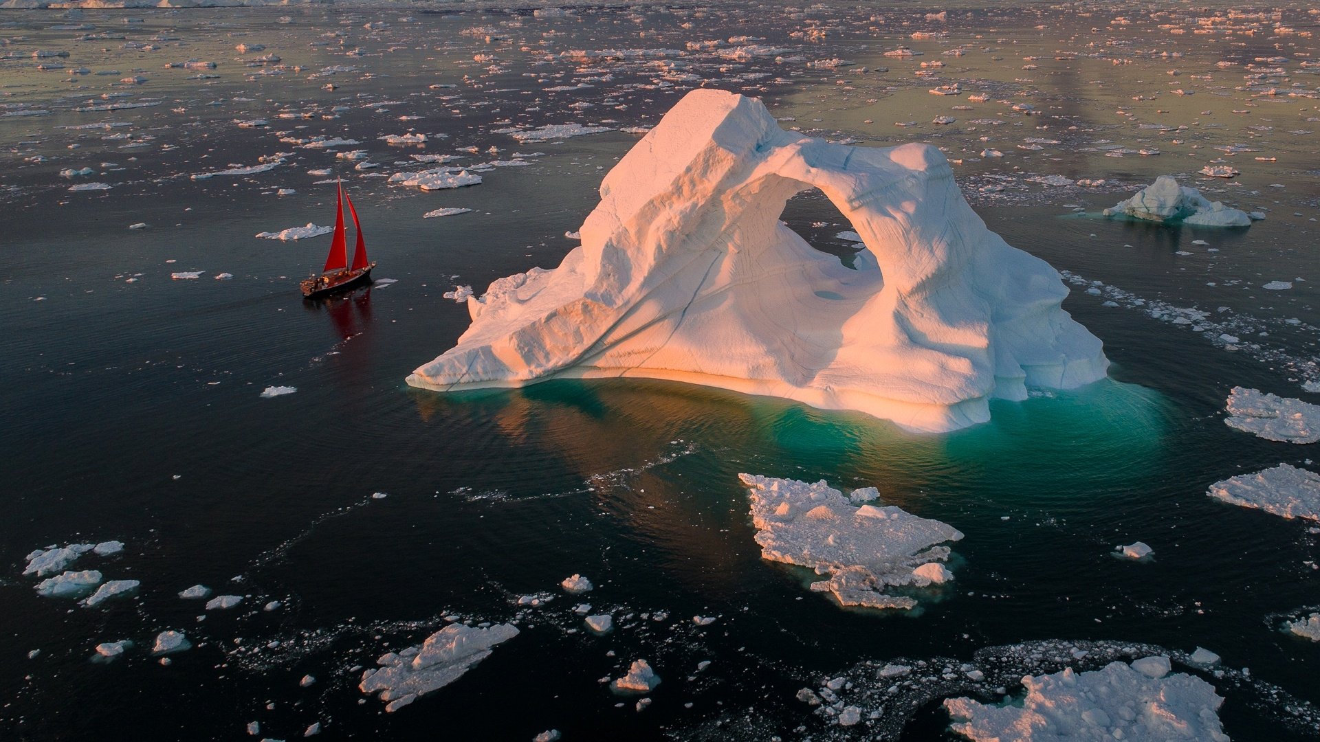 Aerial view of a large iceberg with a natural arch surrounded by floating ice, accompanied by a red sailboat gliding through icy waters in this HD desktop wallpaper.