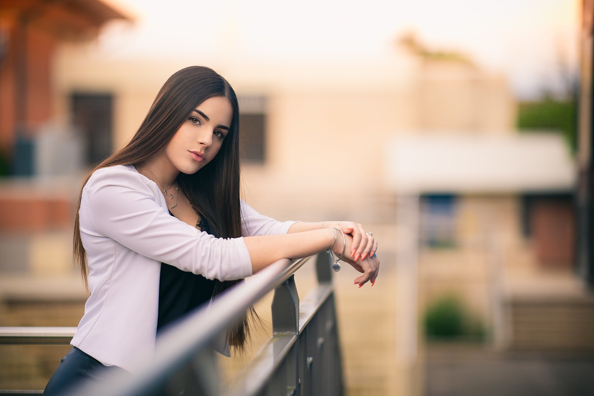 HD PC desktop wallpaper: woman model with black long hair and hazel eyes leaning on a railing, soft depth-of-field urban background.