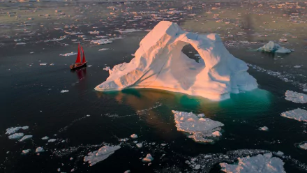 Aerial view of a large iceberg with a natural arch surrounded by floating ice, accompanied by a red sailboat gliding through icy waters in this HD desktop wallpaper.