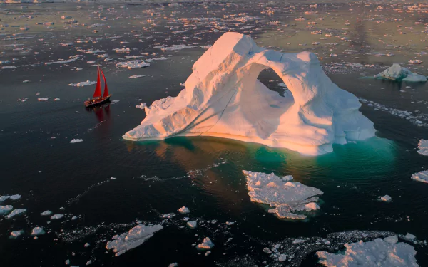Aerial view of a large iceberg with a natural arch surrounded by floating ice, accompanied by a red sailboat gliding through icy waters in this HD desktop wallpaper.
