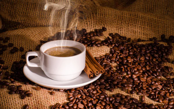 Steaming cup of coffee on a white saucer surrounded by cinnamon sticks and scattered coffee beans on burlap, captured in 4K Ultra HD quality.