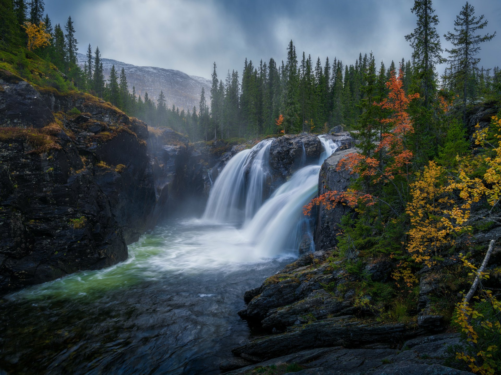 HD desktop wallpaper featuring a stunning nature scene with a flowing river and majestic waterfall, surrounded by vibrant autumn foliage and tall evergreen trees.