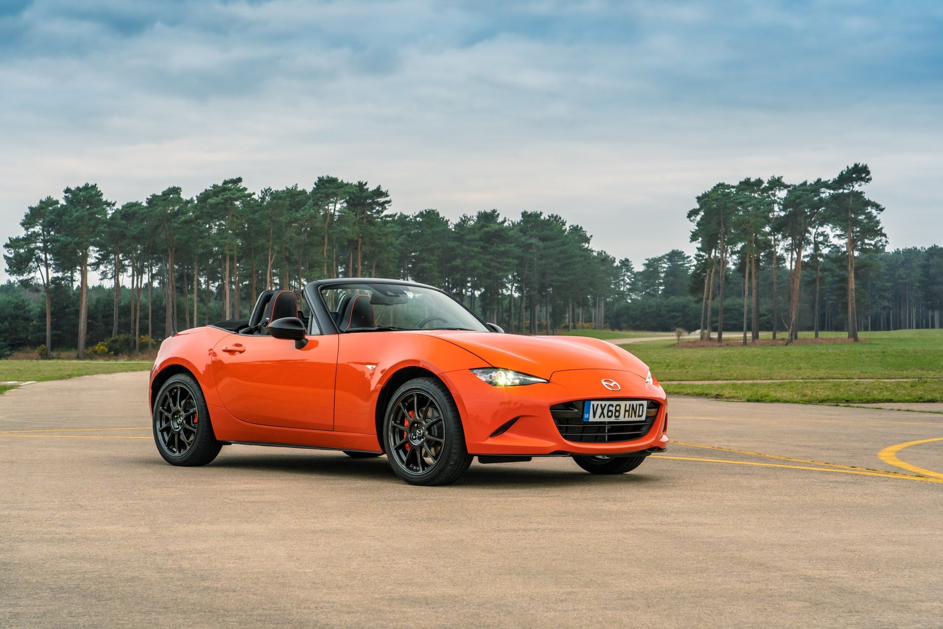 Orange Mazda MX-5 convertible sports car parked on a tarmac with trees and a cloudy sky — 5K Ultra HD PC desktop wallpaper background.