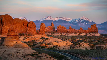 Sunset view of red rock formations and a winding road in Arches National Park, Utah, USA, with snow-capped mountains in the background. 4K Ultra HD landscape wallpaper.