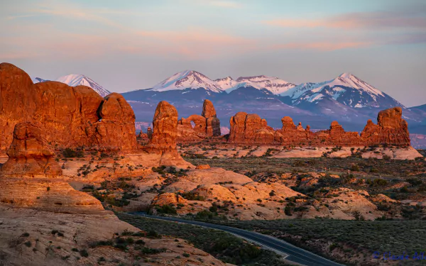 Sunset view of red rock formations and a winding road in Arches National Park, Utah, USA, with snow-capped mountains in the background. 4K Ultra HD landscape wallpaper.