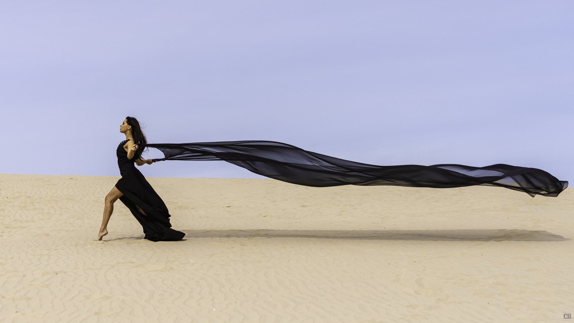 HD PC desktop wallpaper of a woman in a flowing black dress on sand, long sheer train billowing behind her, creating a dramatic, contemplative mood.
