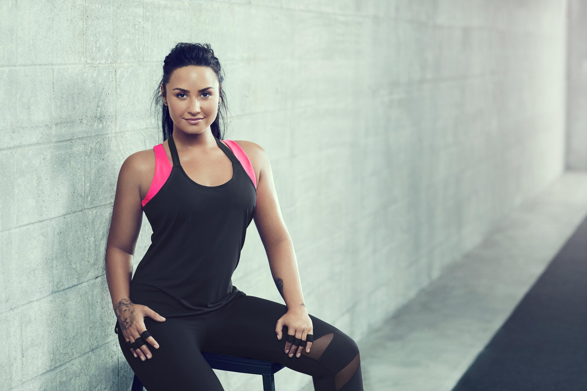 Demi Lovato, American singer with black hair and tattoos, poses confidently in fitness attire against a minimalist concrete background in this 8K Ultra HD wallpaper.