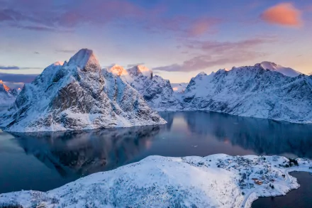 Winter scene of a snowy bay in Lofoten, Norway, with rugged mountains reflected in calm waters under a colorful sky. HD desktop wallpaper photography.