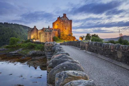 Eilean Donan Castle in Scotland illuminated at dusk, viewed from a stone bridge over calm waters, captured as an HD PC desktop wallpaper and background.