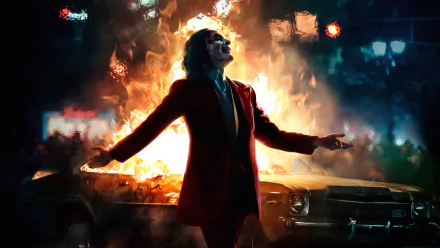 HD desktop wallpaper featuring Joaquin Phoenix as the Joker from the DC Comics movie, standing in front of a burning car with arms outstretched.