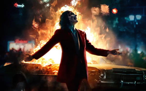 HD desktop wallpaper featuring Joaquin Phoenix as the Joker from the DC Comics movie, standing in front of a burning car with arms outstretched.
