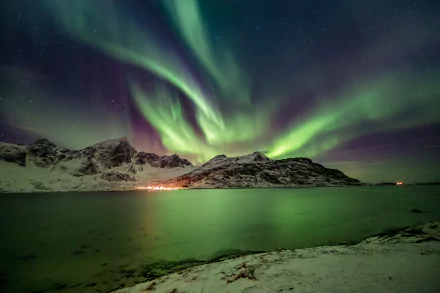 Vibrant aurora borealis lights up a snowy Norwegian landscape on a clear winter night, reflected over calm waters under a starry sky.