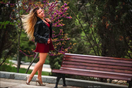 A smiling blonde model with long hair wearing a leather jacket, red dress, and high heels poses beside a wooden bench in a scenic park, captured in HD.