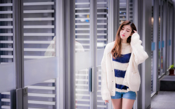 Brunette Asian woman in a striped sweater and shorts poses thoughtfully against a modern glass and metal urban backdrop, captured in 4K Ultra HD.