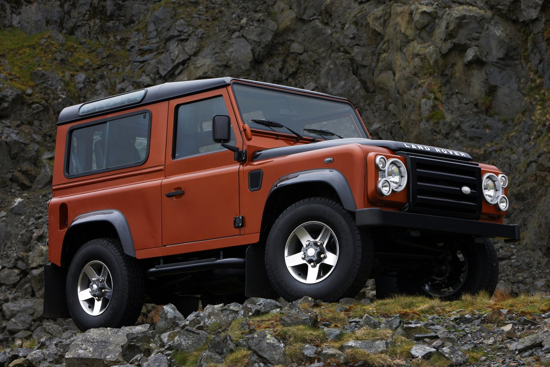 Orange Land Rover Defender SUV parked on rocky terrain, showcased in a sharp 4K Ultra HD desktop wallpaper background.
