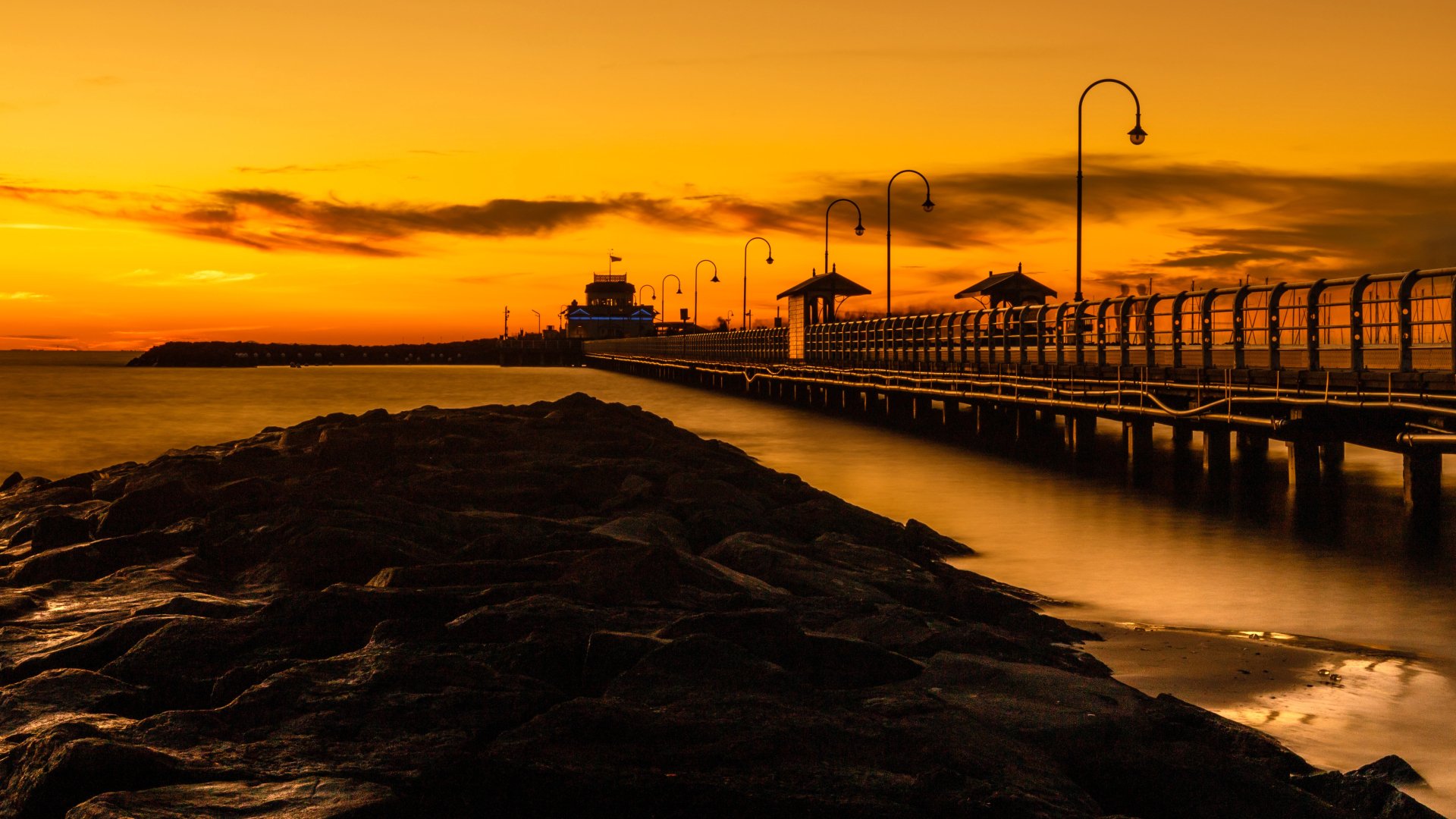 A man-made pier extends into calm waters under a vibrant golden sunset, captured in stunning 4K Ultra HD for a PC desktop wallpaper.