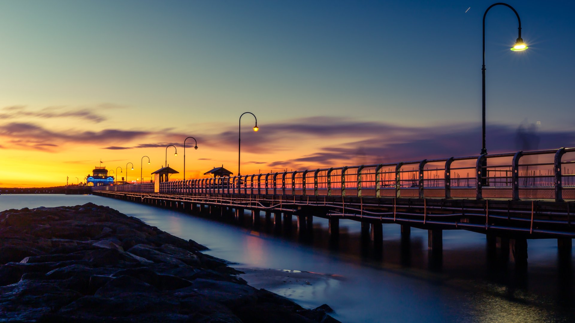 5K Ultra HD PC desktop wallpaper and background: man-made pier lined with lampposts stretching into calm water at sunset, vivid sky and glowing reflections.
