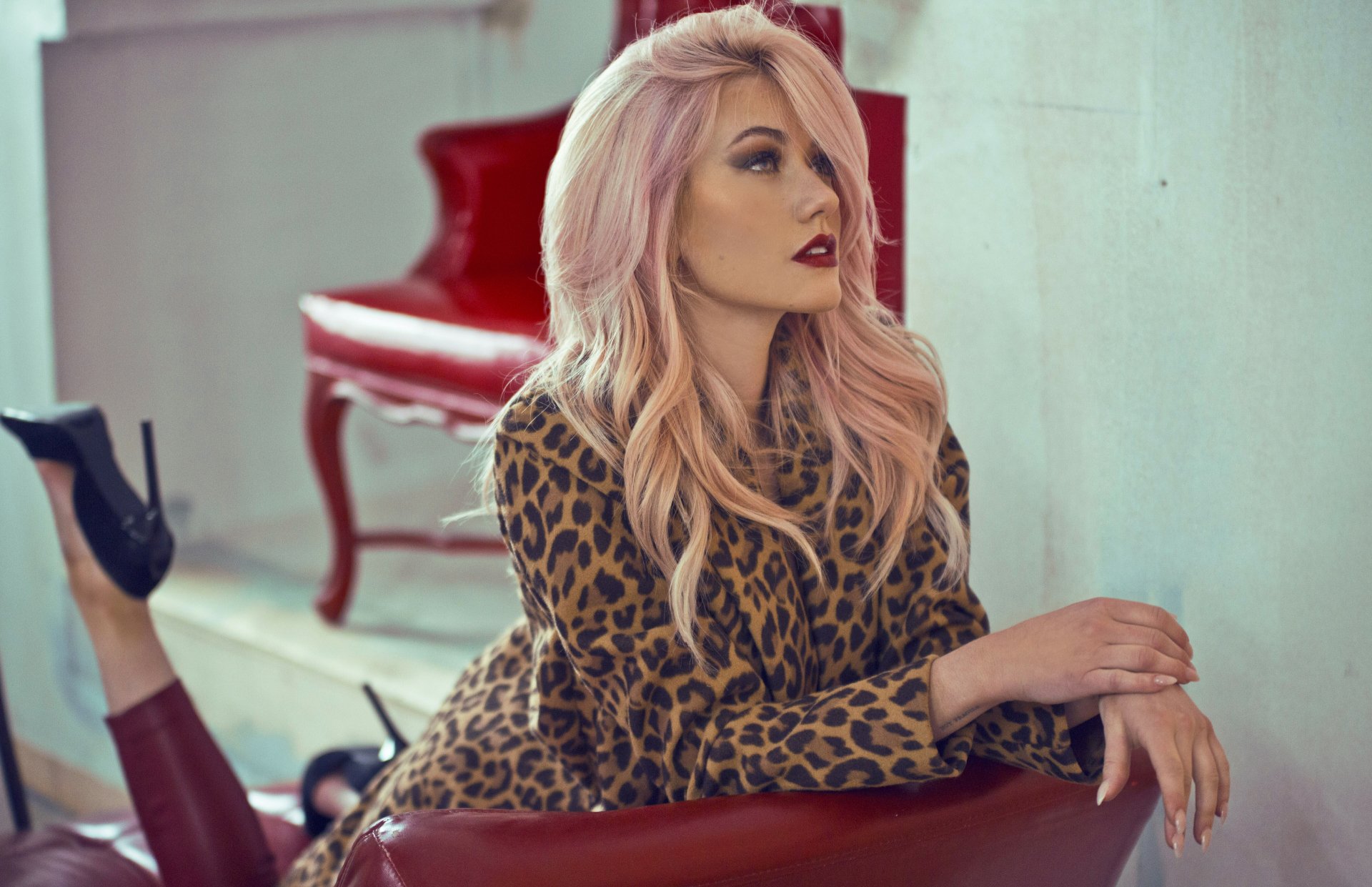 4K Ultra HD wallpaper of American actress Katherine McNamara with pink hair and bold lipstick, lounging on a red leather couch in a leopard print outfit.