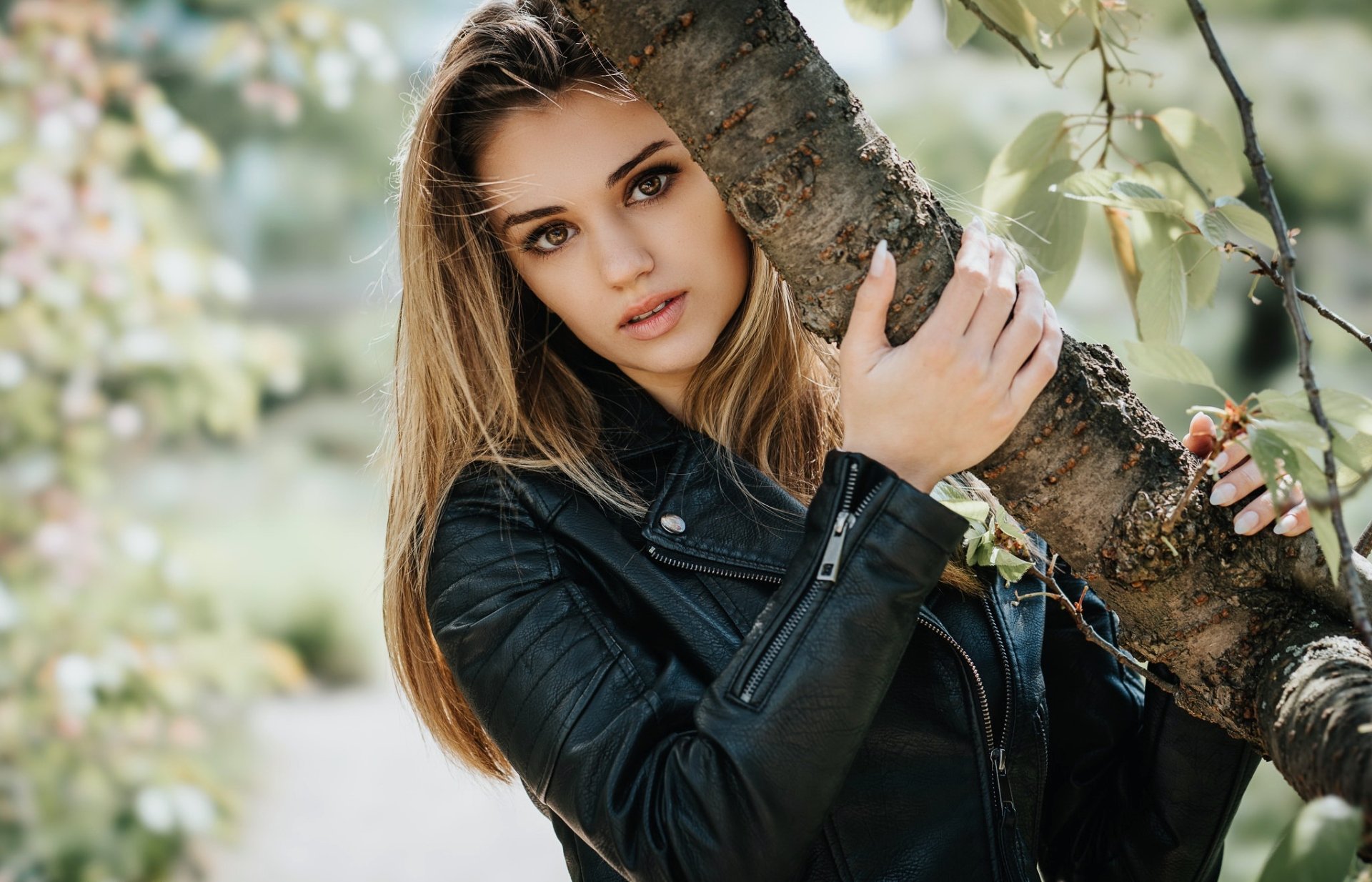 HD desktop wallpaper featuring a woman model wearing a black leather jacket, with a sharp depth of field focusing on her face and the tree branch she holds.