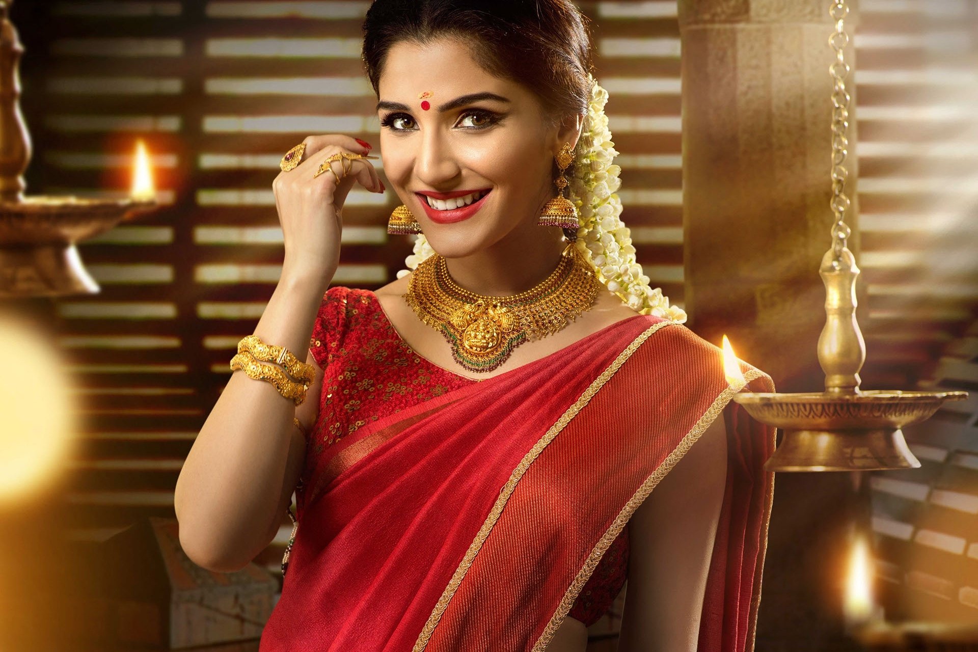 HD PC wallpaper: smiling Indian woman in a red saree with gold necklace, brown eyes and lipstick, jasmine flowers in her hair, warm lamp-lit background.