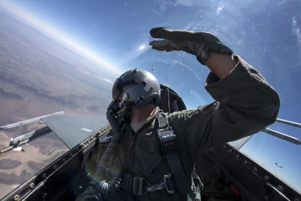 Pilot in a General Dynamics F-16 Fighting Falcon cockpit salutes above a desert; dynamic 5K Ultra HD desktop wallpaper of a military jet and pilot in flight.