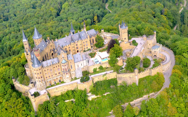 Aerial view of Hohenzollern Castle perched on a forested hilltop in Germany, 4K Ultra HD desktop wallpaper showing the man-made fairy-tale fortress and surrounding green landscape.