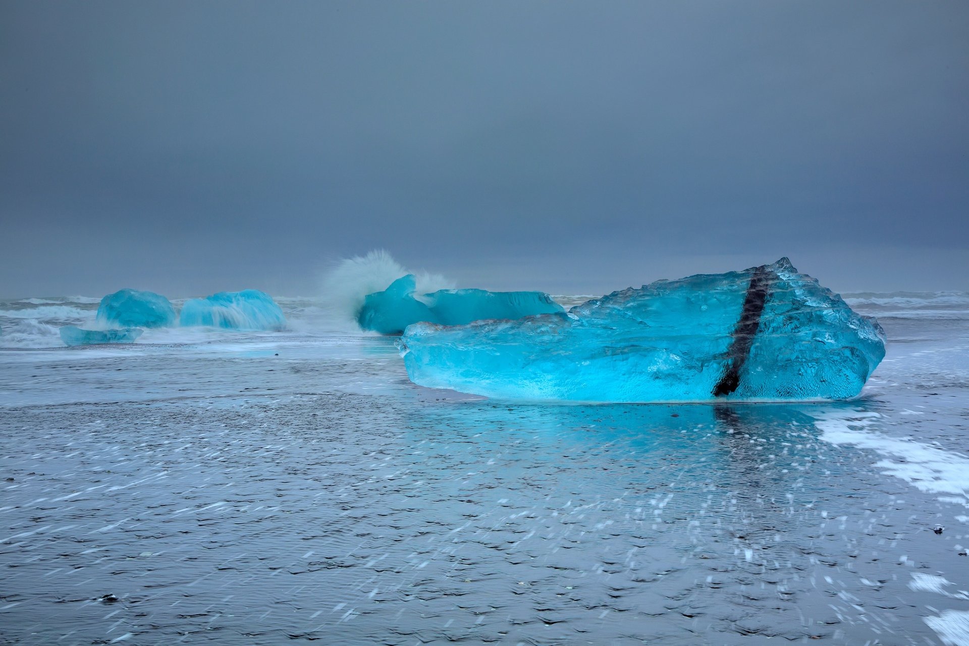 HD PC desktop wallpaper: nature scene of vivid blue icebergs and ice chunks on a reflective, icy shoreline beneath a muted, overcast sky.