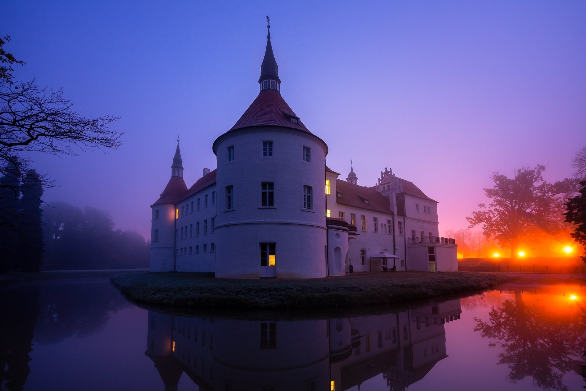 A man-made castle surrounded by a moat reflects in still water under a vibrant night sky, bathed in soft purple and orange hues.