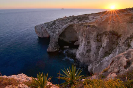 4K Ultra HD PC desktop wallpaper of Malta coastline at sunset: rugged sea cliffs and arches over deep blue water, coastal plants in foreground, warm sunburst on the horizon.