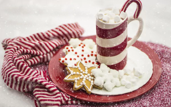 HD desktop wallpaper of a festive scene with a striped mug of hot chocolate, a candy cane, and assorted holiday cookies on a red plate, set against a snowy background. Christmas decorations enhance the holiday spirit.