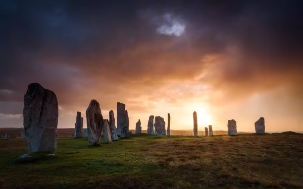 HD wallpaper of the Callanish standing stones in Scotland, set against a dramatic sunset sky with natural landscape.