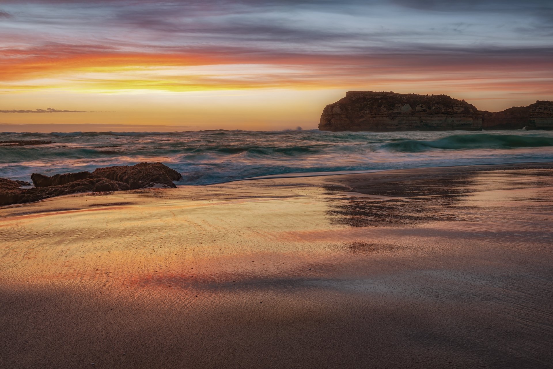 4K Ultra HD desktop wallpaper showcasing a serene beach at sunset with gentle ocean waves and a rocky cliff under a colorful sky.