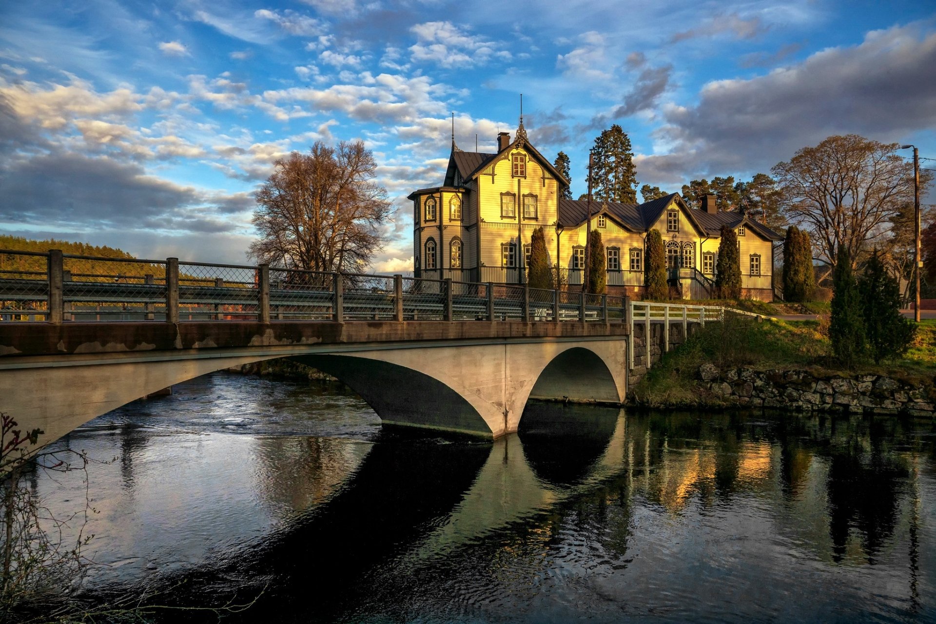 HD desktop wallpaper of a man-made bridge spanning a river in Kouvola, Finland, with historic buildings and a partly cloudy sky in the background.