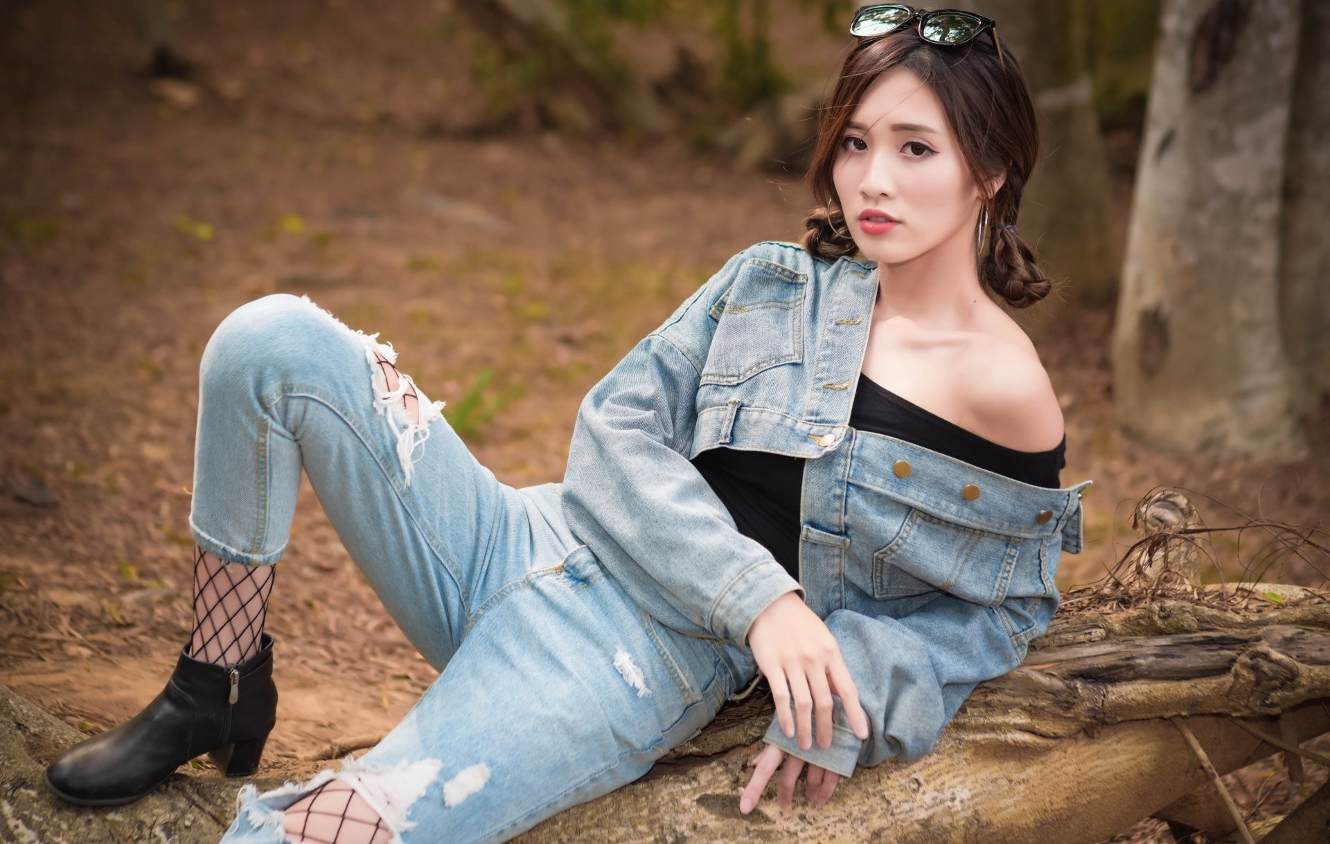 Asian brunette woman wearing denim jacket and jeans with sunglasses on her head, posing outdoors on a fallen tree.