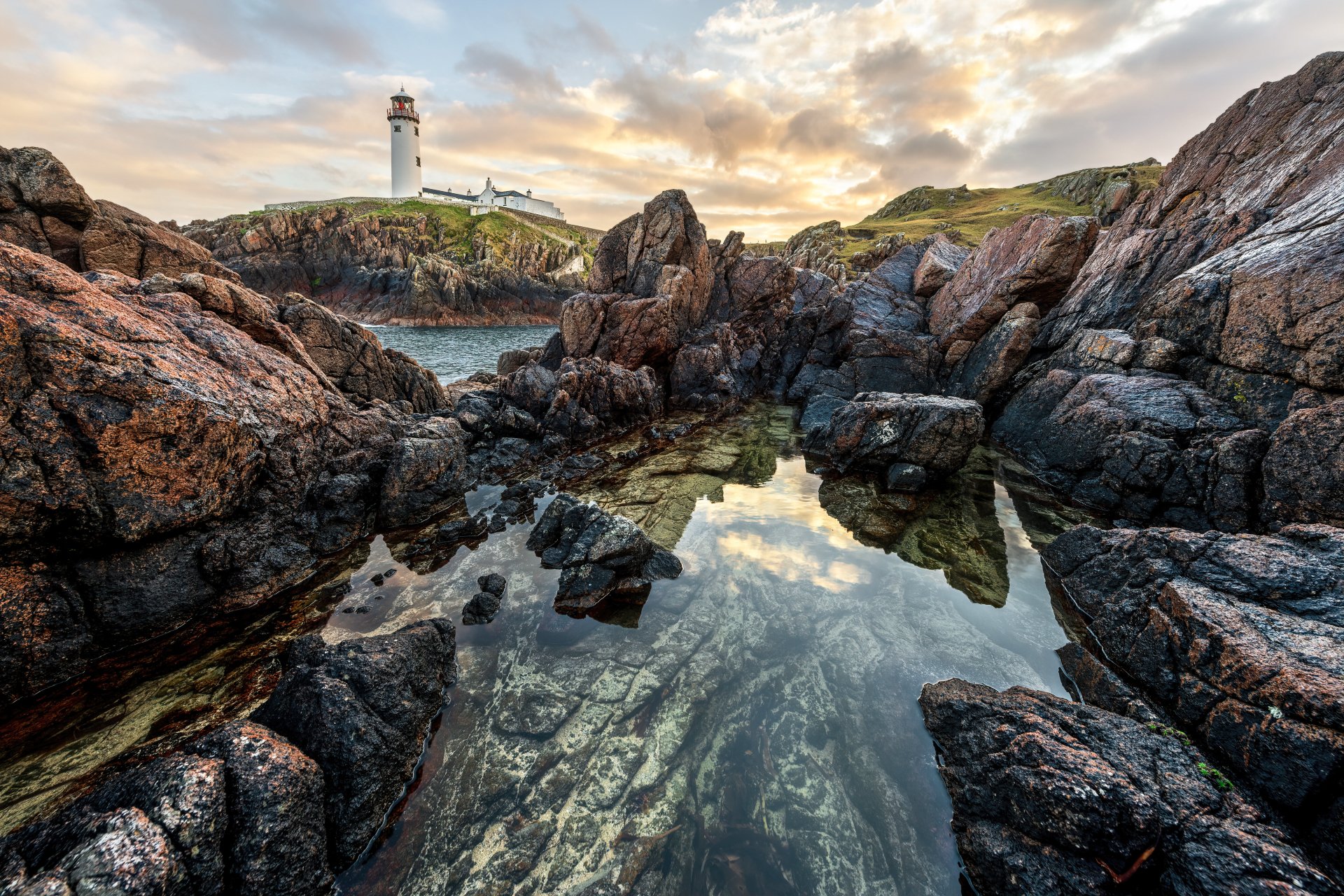 5K Ultra HD PC desktop wallpaper: Irish coastline with a man-made lighthouse on a rocky headland, sunset sky reflected in a tidal pool among jagged rocks.