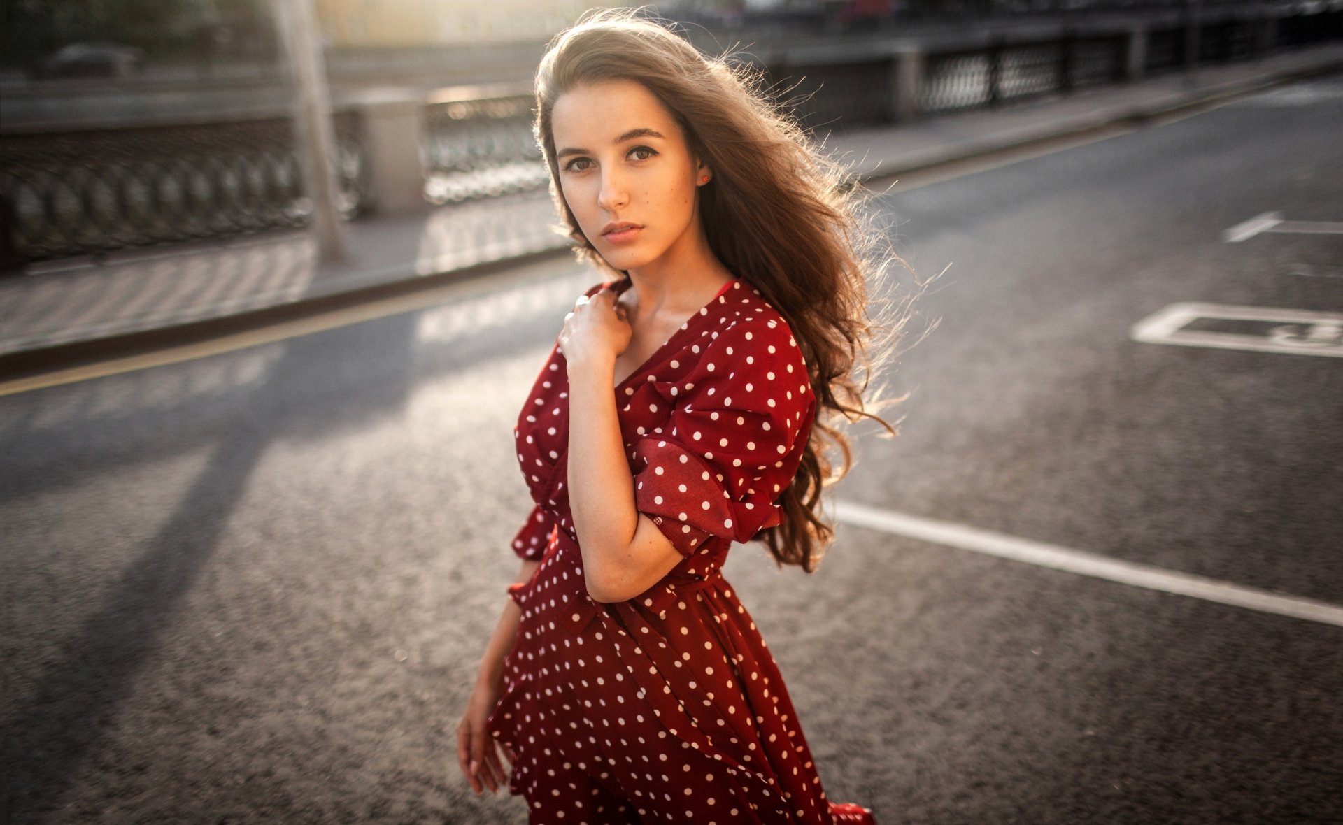 2K Quad HD PC desktop wallpaper: brunette model with long hair in a red polka-dot dress standing on an urban street, backlit by warm evening light, elegant background