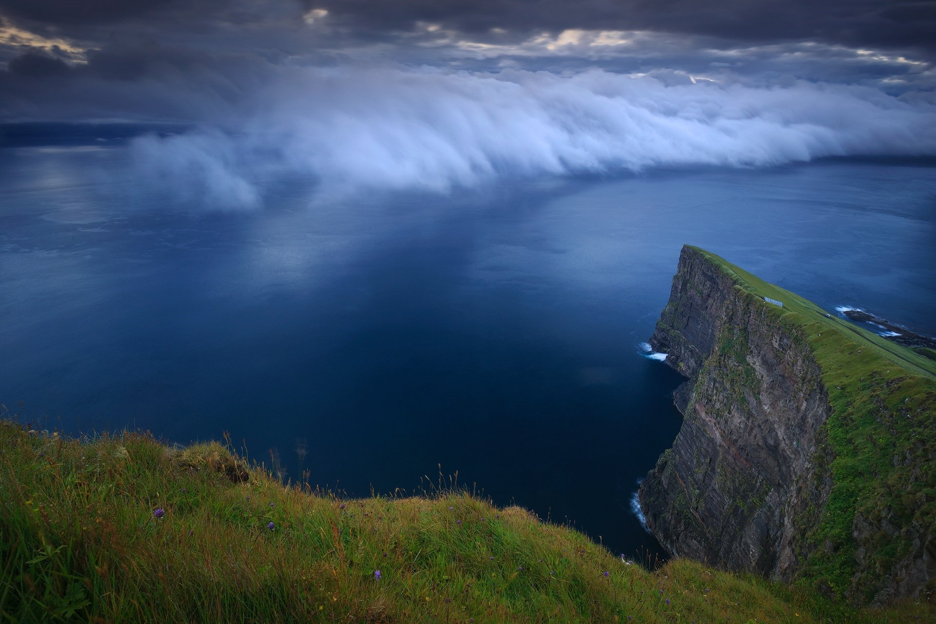Faroe Islands coastal cliff rising above a deep blue ocean under rolling clouds, with lush grassy foreground — HD PC desktop wallpaper background.