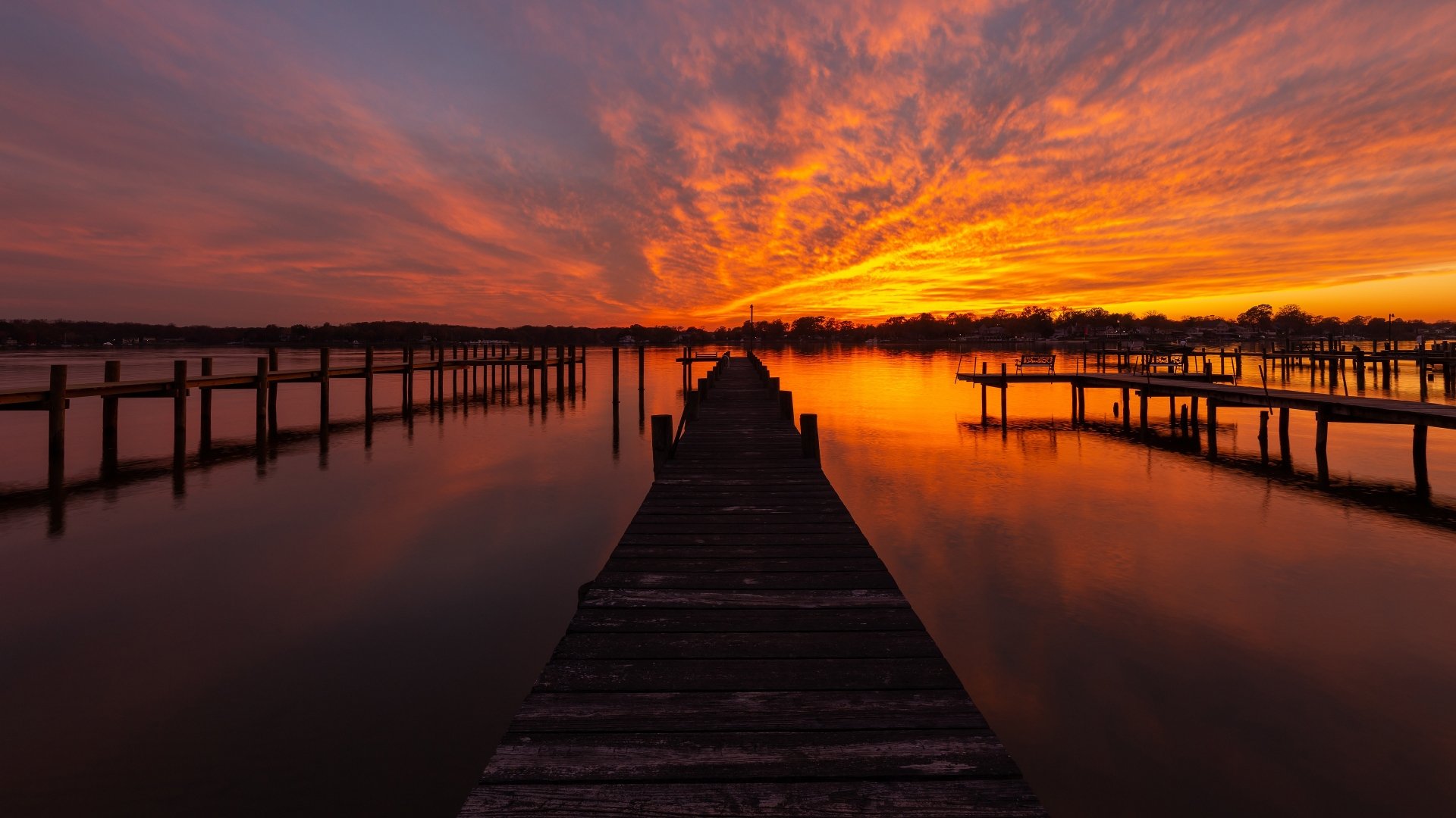 4K Ultra HD PC desktop wallpaper: wooden pier at twilight stretching into glassy water, fiery evening sunset sky reflected across the lake, man-made docks silhouetted along the shore.