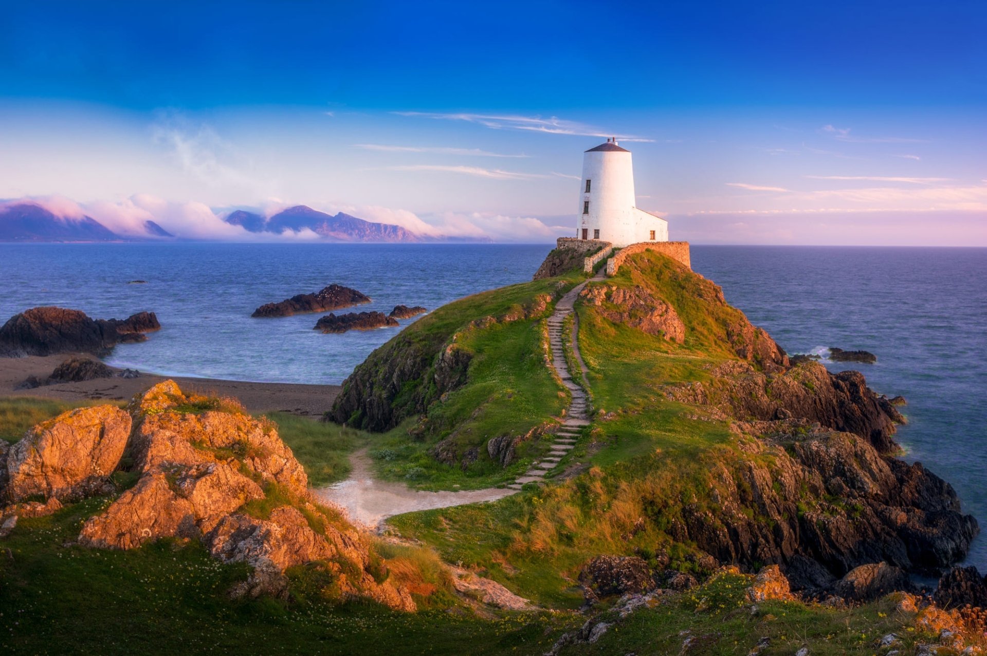 HD PC desktop wallpaper and background showing a white man-made lighthouse atop a green rocky island with a winding stone path to the sea under a colorful sunset.