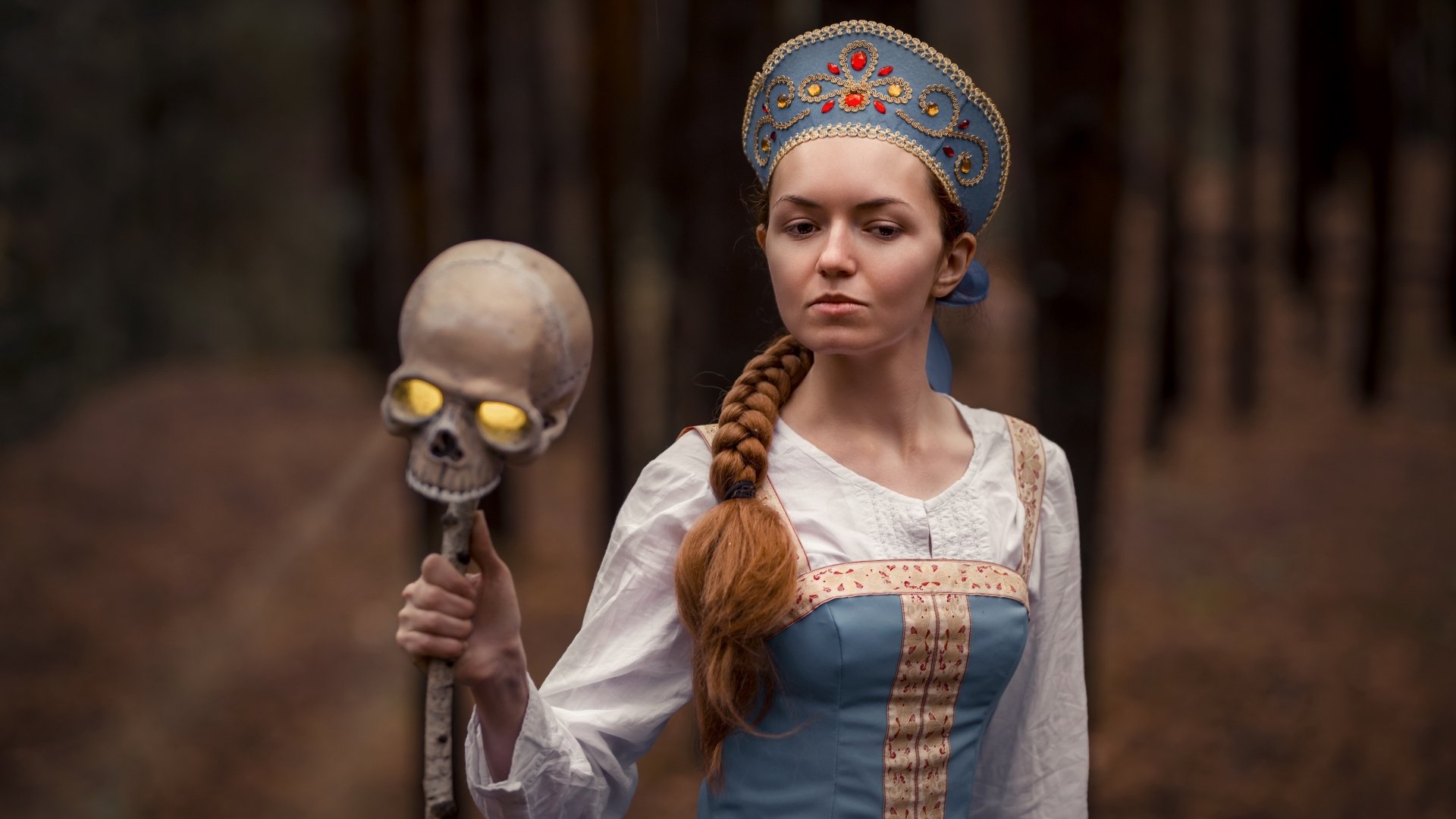 A woman in detailed witch cosplay holds a skull staff, set against a blurred forest background, captured in 4K Ultra HD as a PC desktop wallpaper.