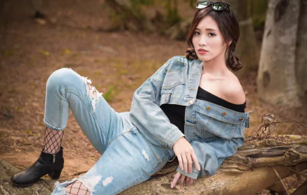 Asian brunette woman wearing denim jacket and jeans with sunglasses on her head, posing outdoors on a fallen tree.